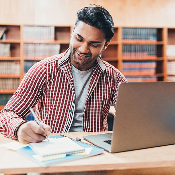 A young man writing on a notepad with his laptop open while in the school library.